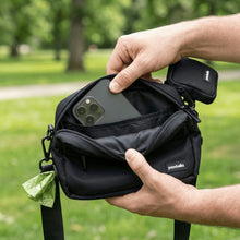 Outdoors in an Australian park with green grass and trees, a person puts a smartphone into the front pocket of a small black Pawbella Crossbody Dog Walking Bag with a pawbell tag.