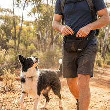A person wearing shorts and the Pawbella Dog Walking Bum Bag walks on a dirt trail in the Australian woods, accompanied by a black and white dog that looks up at them attentively.